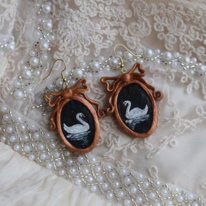 May include: A pair of handmade earrings featuring a swan painting within a copper-colored frame. The oval frames have a bow at the top and are suspended from gold-colored ear wires. The earrings rest on a cream-colored, lace-textured surface, with a pearl necklace.