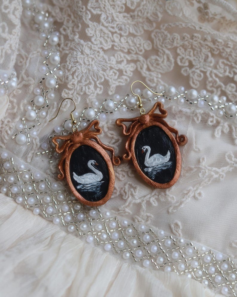 May include: A pair of handmade earrings featuring a swan painting within a copper-colored frame. The oval frames have a bow at the top and are suspended from gold-colored ear wires. The earrings rest on a cream-colored, lace-textured surface, with a pearl necklace.