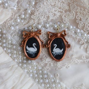 May include: A pair of handcrafted earrings featuring oval frames with a copper-colored bow design. Each frame holds a miniature painting of a white swan on a black background. The earrings have gold-colored hooks.