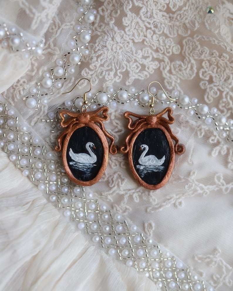 May include: A pair of handcrafted earrings featuring oval frames with a copper-colored bow design. Each frame holds a miniature painting of a white swan on a black background. The earrings have gold-colored hooks.
