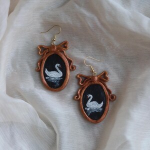 May include: A pair of handcrafted earrings featuring a swan painting within a bronze-colored frame. The oval frames are adorned with a bow at the top and hang from gold-colored ear wires. The swan is white, set against a black background.