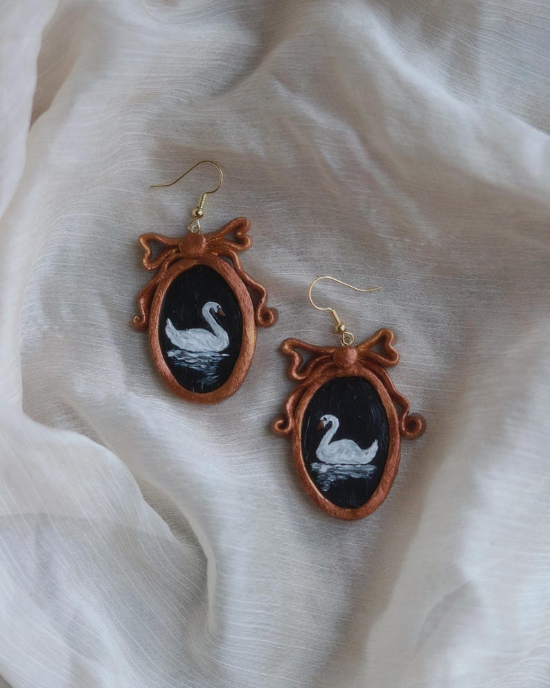 May include: A pair of handcrafted earrings featuring a swan painting within a bronze-colored frame. The oval frames are adorned with a bow at the top and hang from gold-colored ear wires. The swan is white, set against a black background.