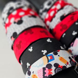 May include: A collection of fabric hair scrunchies featuring various patterns including red with white polka dots, solid red, black with white mouse head silhouettes, and a white print with colorful mouse head icons, cupcakes, and ice cream treats.
