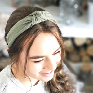 May include: A green knotted headband made of a soft, ribbed fabric. The headband is worn by a person with long brown hair.