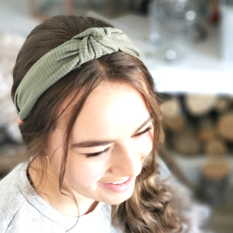 May include: A green knotted headband made of a soft, ribbed fabric. The headband is worn by a person with long brown hair.