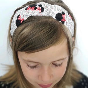 May include: A white fabric headband featuring a repeating pattern of black Minnie Mouse silhouettes with red and white polka dot bows, interspersed with the text Minnie Mouse printed in a grey font across the entire surface of the accessory.