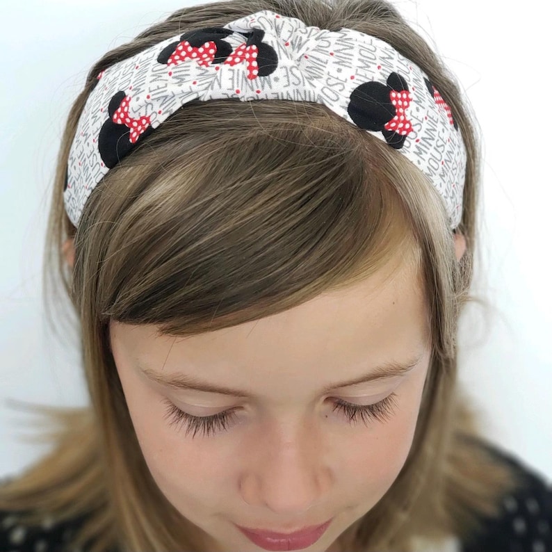 May include: A white fabric headband featuring a repeating pattern of black Minnie Mouse silhouettes with red and white polka dot bows, interspersed with the text Minnie Mouse printed in a grey font across the entire surface of the accessory.