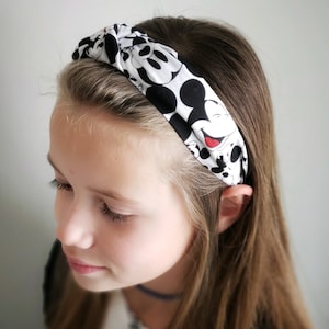 May include: A knotted fabric headband featuring a repeating black and white pattern of Mickey Mouse faces with small red accents on the mouth. The headband is made of a soft material and has a twisted knot detail at the top center of the band.