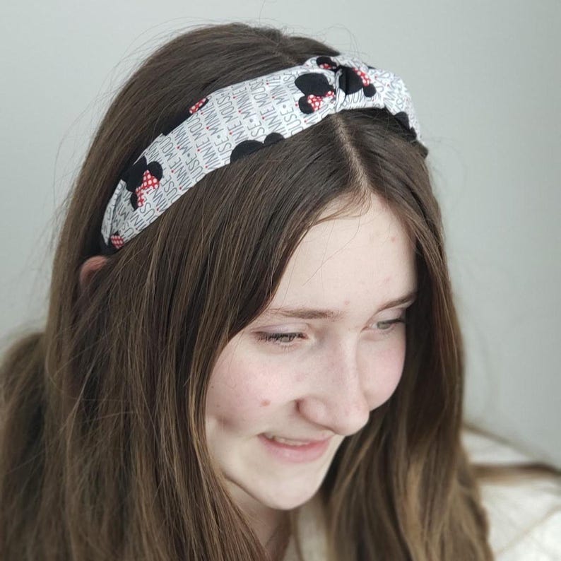 May include: A knotted fabric headband featuring a repeating pattern of black and white Minnie Mouse silhouettes with red polka dot bows, interspersed with the text "MINNIE MOUSE" printed in a black sans-serif font against a white background.