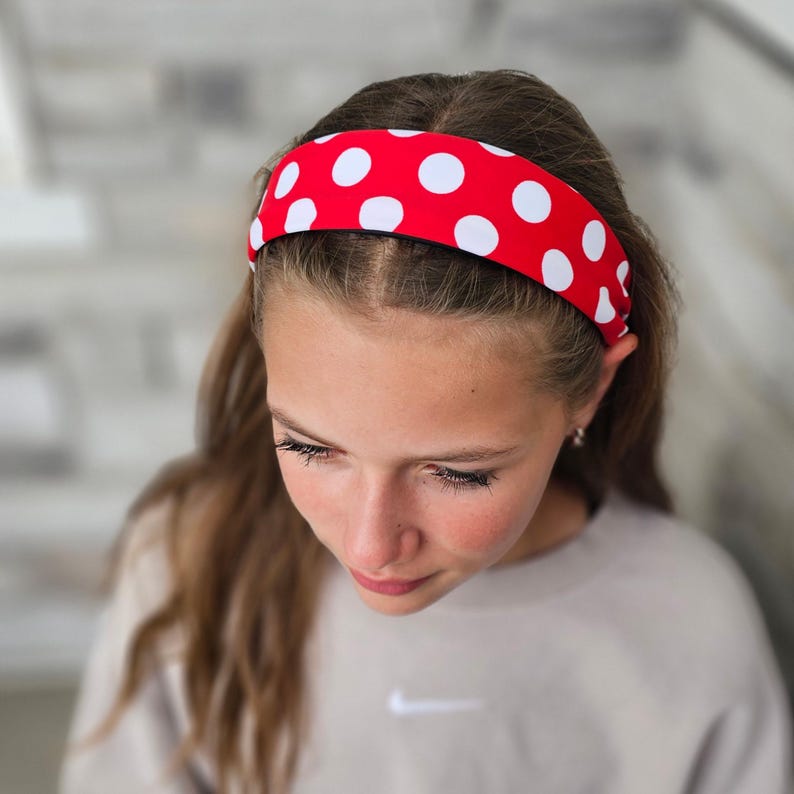 May include: A wide headband featuring a vibrant red base color with a repeating pattern of large, circular white polka dots. The fabric accessory is positioned across the top of the head, securing hair away from the face with a smooth, consistent fit.