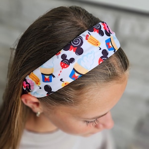 May include: A white fabric headband featuring a repeating pattern of black mouse-ear silhouettes, light blue soft-serve ice cream cones, yellow popsicles, and red accents. The design includes various colorful snack-themed icons on a white background.