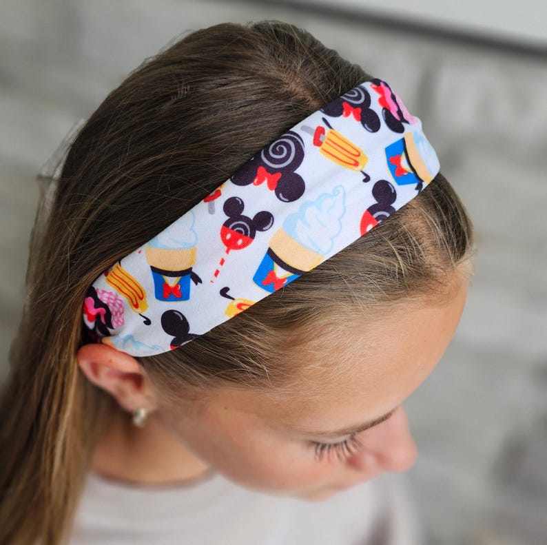 May include: A white fabric headband featuring a repeating pattern of black mouse-ear silhouettes, light blue soft-serve ice cream cones, yellow popsicles, and red accents. The design includes various colorful snack-themed icons on a white background.