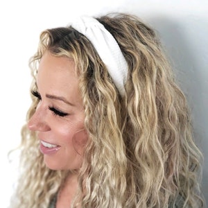 May include: A white knotted headband made of a soft, textured fabric. The headband is worn on a person with long, curly blonde hair.