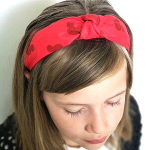 May include: A bright red fabric headband featuring a knotted center detail and a repeating dark red silhouette pattern of mouse ears. The headband is constructed from a soft material and sits across the top of the head with a wide band design.