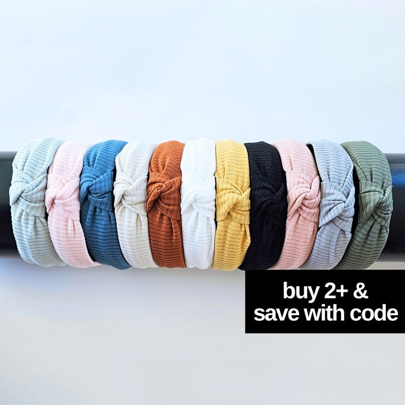 May include: A set of 10 knotted headbands in various colors, including light blue, pink, white, orange, yellow, black, and green. The headbands are arranged in a row on a black surface. The text "buy 2+ & save with code" is visible below the headbands.