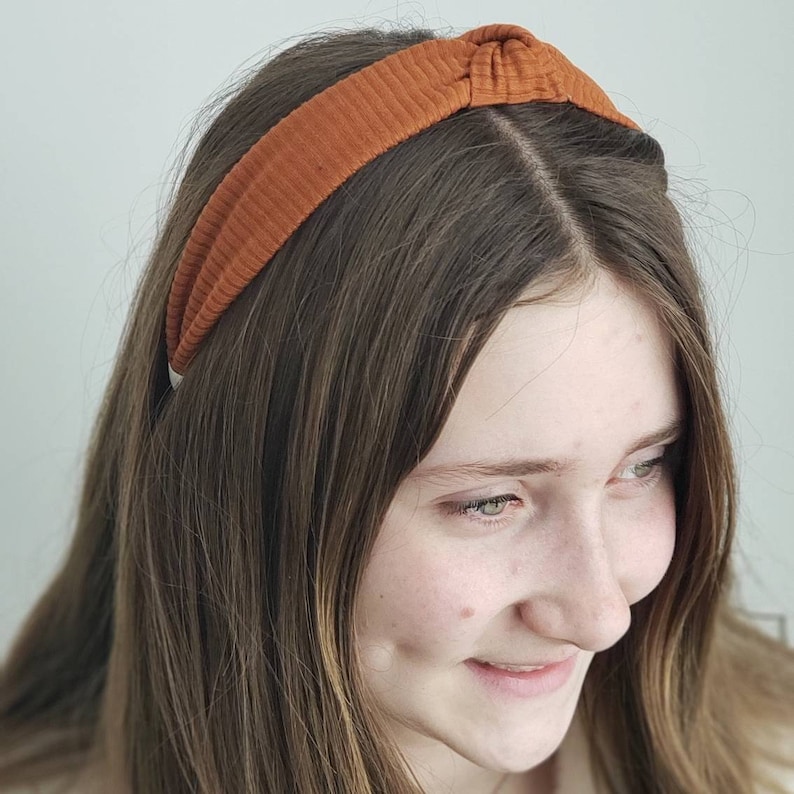 May include: A brown fabric headband with a knotted detail on top. The headband is worn by a person with long brown hair.
