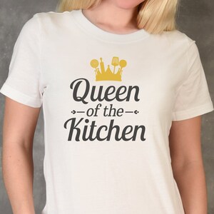 May include: White t-shirt with the text "Queen of the Kitchen" in black script, topped with a gold crown and kitchen utensil graphic. The shirt is short-sleeved and made of a soft material.