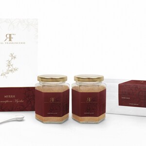 May include: Two glass jars with gold lids filled with brown powder labeled "Myrrh Commiphora Myrrha" and "Royal Frankincense". The jars are on a white surface with a white box and a white scoop.