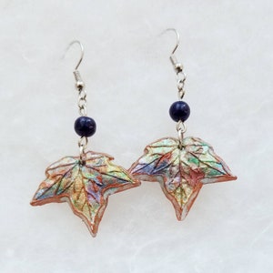 May include: A pair of leaf-shaped earrings. Each earring features a detailed, multi-colored leaf pendant with shades of red, green, and gold. A dark blue bead hangs above each leaf, connected by a silver chain and hook.