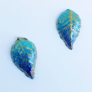 May include: Two leaf-shaped decorative objects. Each leaf has a blue and gold color scheme. The leaves have a textured surface and are painted with shades of blue and gold. The leaves are on a white background.