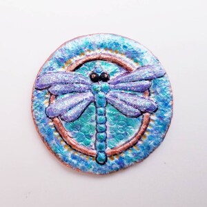 May include: A blue and purple dragonfly with glitter accents is painted on a round, blue and gold background. The dragonfly is in a circle with a copper border.