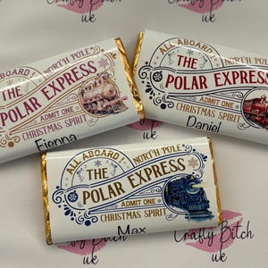 Pode incluir: Três barras de chocolate embaladas com bordas de folha de ouro. Cada barra apresenta um design "Polar Express" com uma ilustração de trem e os nomes "Fienna", "Daniel" e "Max". As barras são brancas com detalhes em vermelho e azul.