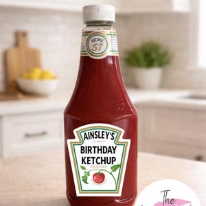 May include: A bottle of red ketchup with a custom label that reads "Ainsley's Birthday Ketchup." The label is in a green and white design with a red tomato illustration. The bottle has a white cap and a "Heinz 57" label.