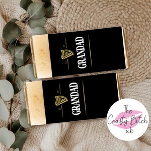 May include: Two chocolate bars with black wrappers featuring the word "GRANDAD" in white, a gold harp logo, and the numbers "17" and "59". The bars have a light-colored top section. The background includes a beige knitted texture and eucalyptus leaves.