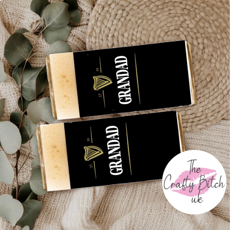 May include: Two chocolate bars with black wrappers featuring the word "GRANDAD" in white, a gold harp logo, and the numbers "17" and "59". The bars have a light-colored top section. The background includes a beige knitted texture and eucalyptus leaves.