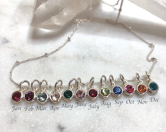 May include: A silver chain necklace with 12 small charms, each featuring a different colored crystal. The charms are labeled with the months of the year, from January to December.