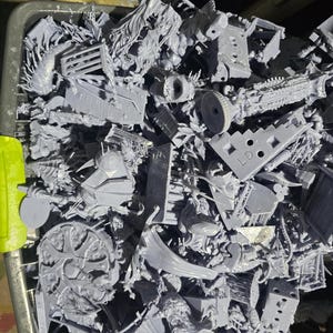 May include: A bin filled with numerous small, gray plastic model parts. The parts vary in shape and size, including what appear to be architectural elements, mechanical components, and abstract shapes. The models are likely for miniature wargaming or model building.