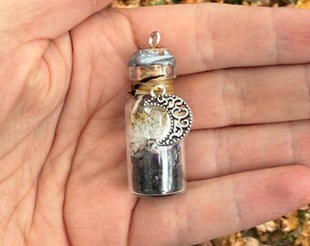 May include: A small glass vial filled with crystals and stones, topped with a silver and gray stopper. A silver crescent moon charm hangs from the vial. The vial is held in a hand against a backdrop of fall leaves.