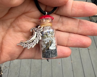 May include: A small glass bottle pendant necklace filled with various natural elements. The bottle has a red cap and a silver-toned charm with a crescent moon and wing design. The necklace cord is black.