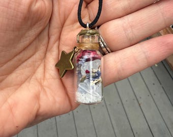May include: A small glass bottle pendant filled with colourful crystals and dried flowers, suspended from a black cord. A gold-coloured star charm hangs from the bottle. The bottle is sealed with a cork and twine.