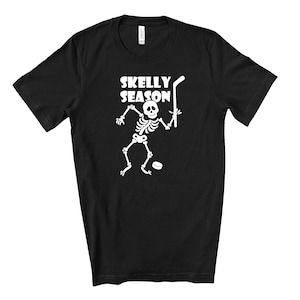 May include: Black t-shirt with the words "SKELLY SEASON" above a white skeleton playing hockey. The skeleton is holding a hockey stick and a puck is on the ground. The shirt is made of a soft material.