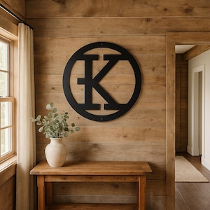 May include: A black metal wall art piece featuring the letter "K" inside a circular frame. The artwork is mounted on a wooden wall above a wooden table. A vase with greenery sits on the table.