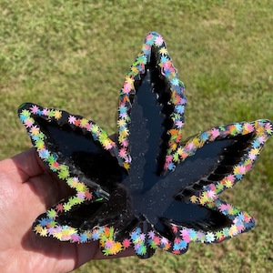 May include: Black resin ashtray in the shape of a cannabis leaf, with colorful star-shaped glitter around the edges. The ashtray is held in a hand against a background of green grass.