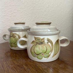 May include: Two ceramic lidded bowls with handles. The bowls are off-white with a green and brown floral design. The lids are off-white with a brown rim. The bowls are sitting on a wooden surface.