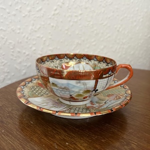 May include: A vintage teacup and saucer set featuring a delicate floral and landscape design in shades of red, gold, and white. The cup and saucer are made of porcelain and have a gold rim.