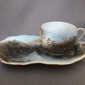 May include: A white porcelain teacup and saucer set featuring a hand-painted landscape scene in shades of blue, brown, and gold. The scene depicts a Japanese-style landscape with trees, a house, and a river.