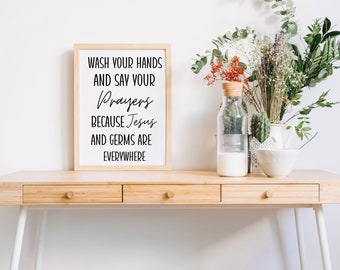 May include: A framed print with the text "Wash your hands and say your prayers because Jesus and germs are everywhere". The print is on a wooden table with a vase of flowers and a cactus.