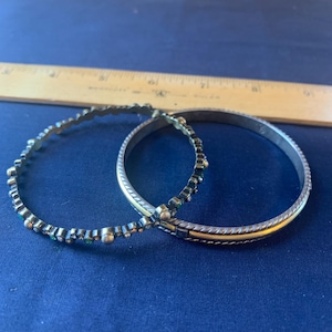 May include: Two silver-toned bangles with decorative detailing. One bangle features small, round embellishments and the other has a textured design with a gold-colored band. The bangles are displayed on a blue surface.