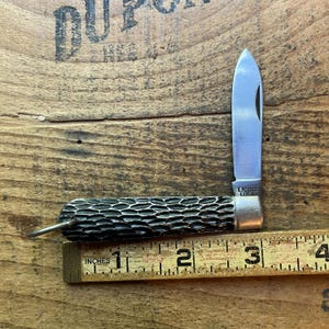 May include: A folding pocket knife with a textured black handle and a single silver-colored blade. The knife is positioned against a yellow measuring tape, showing a length of approximately 3.5 inches from the end of the handle to the blade tip.
