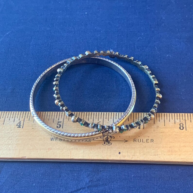 May include: Two silver-toned bangles with decorative detailing. One bangle has a textured rope design, while the other features small, dark-colored embellishments. The bangles are on a blue surface next to a wooden ruler.