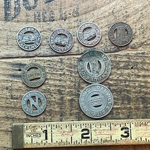May include: A collection of vintage transit tokens, including tokens from Bluebird Coach Lines, Danville City Lines, and SMTD. The tokens are silver-colored and feature various designs, including buses and text. A wooden ruler with inch measurements is in the foreground.