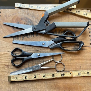 May include: Three pairs of scissors on a wooden surface. The scissors are black and silver with a variety of blade styles. A wooden ruler with inch markings is also visible.