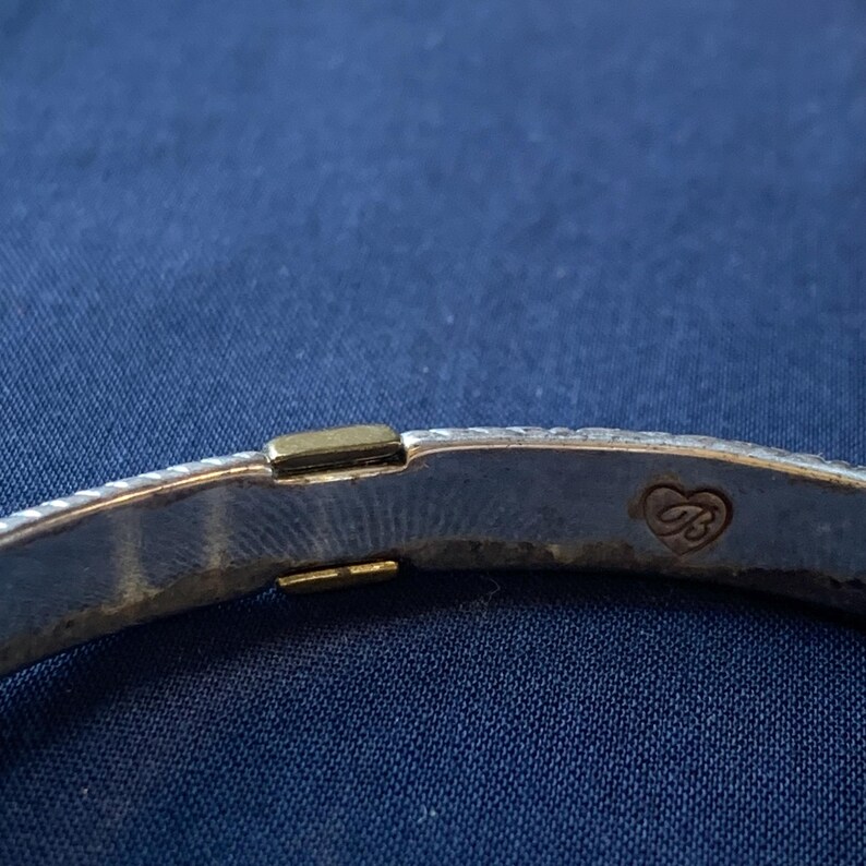 May include: Close-up of a silver-toned bracelet with a textured design. The bracelet features a gold-colored clasp and a heart-shaped emblem with the letter "B". The background is a solid blue fabric.