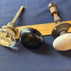 May include: Three door knobs are arranged with a yellow wooden ruler. One knob is clear glass with a fluted design, one is a smooth black sphere, and one is a white ceramic knob with a metal spindle and mounting plate, measuring up to 9 inches.