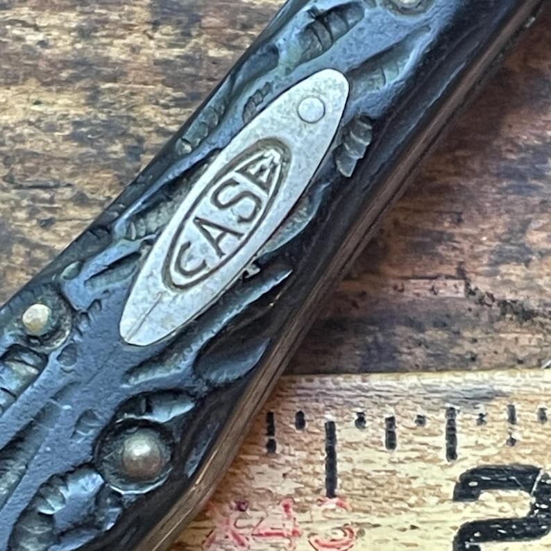 May include: Close-up of a vintage pocket knife with a textured, dark handle. The knife features a silver inlay with the word "CASE" in a curved font. The handle has decorative rivets and a worn appearance, suggesting age and use.