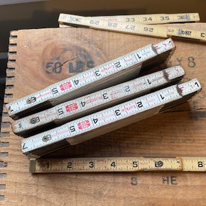May include: Four vintage Lufkin folding rulers with white and black markings. The rulers are made of wood and have red and black lettering. The rulers are stacked on top of each other. The top ruler is partially visible. The rulers are on a wooden surface.